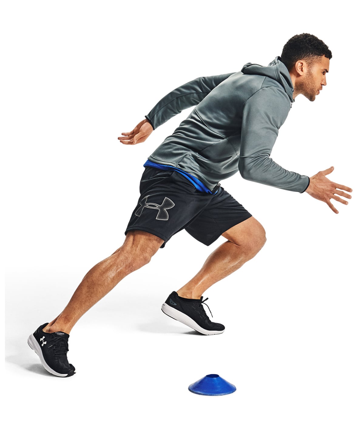 A man in athletic wear, featuring a gray long-sleeve hoodie, black Under Armour shorts, and black running shoes, is captured mid-stride running on a white studio background. A small blue cone is visible on the ground in front of him.