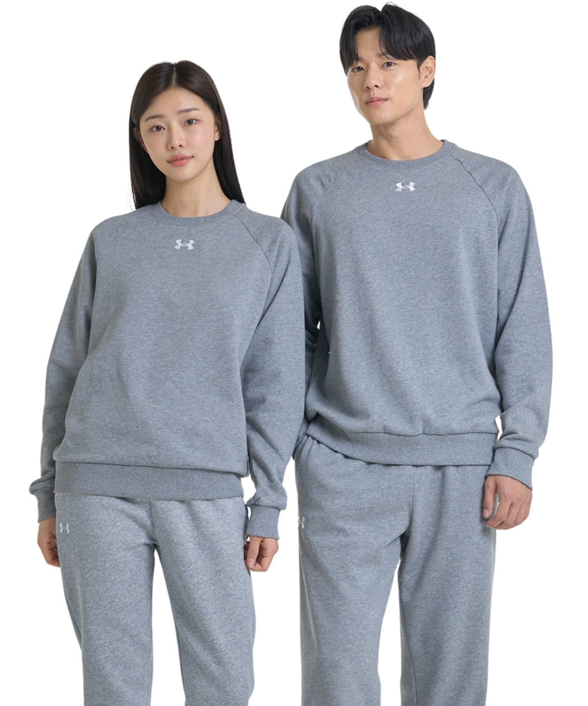 A man and a woman modeling matching gray Under Armour sweatshirt and sweatpants sets against a plain white background.