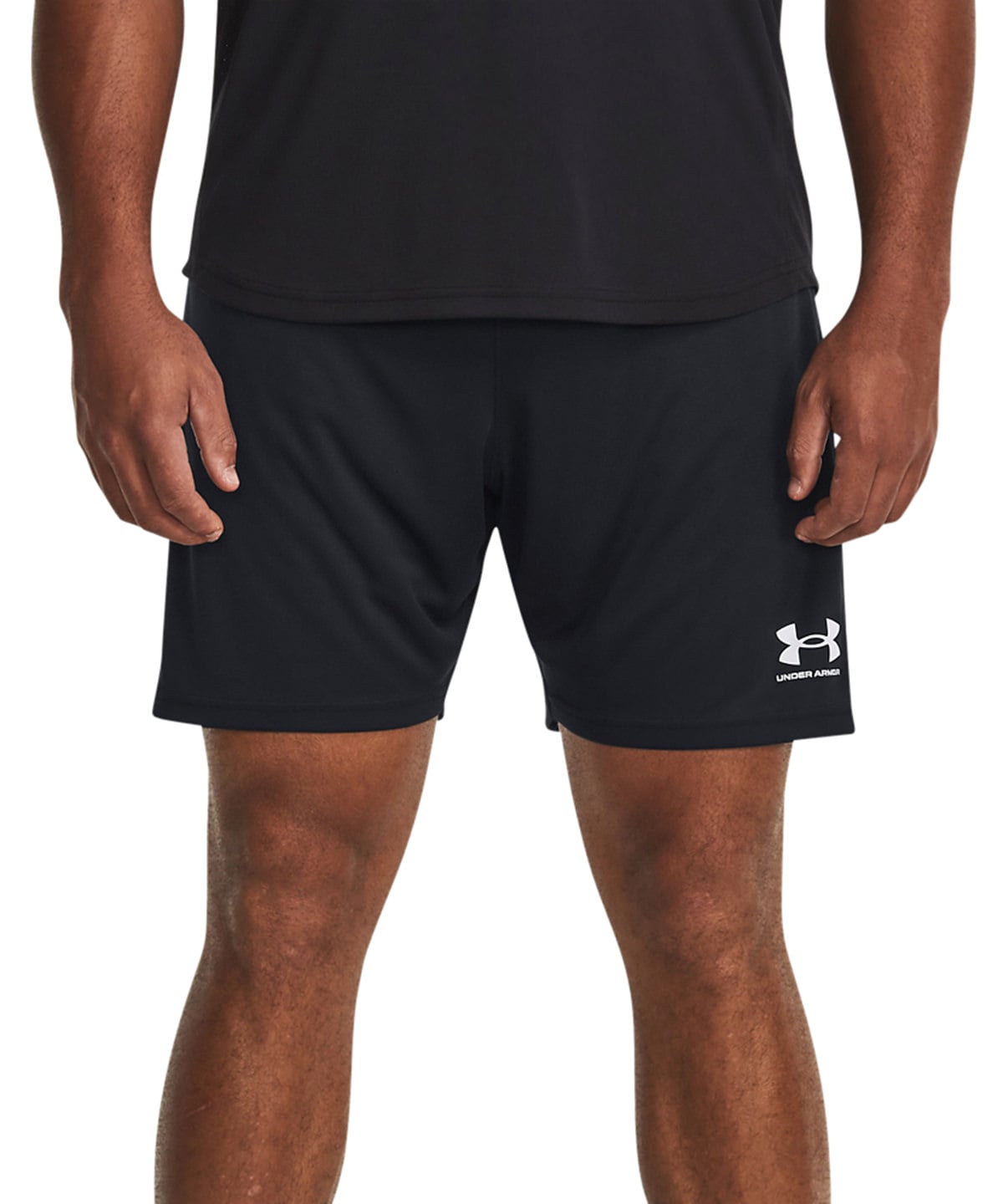 A close-up of a person wearing black athletic shorts with a white Under Armour logo on the lower left leg, paired with a black shirt against a plain white background.